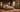A rustic bedroom furniture set in a warm, styled room. The set includes a bed frame with a tall headboard, two matching nightstands, a chest of drawers, and a dresser with a matching mirror.