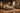 A rustic bedroom furniture set in a warm, styled room. The set includes a bed frame with a tall headboard, two matching nightstands, a chest of drawers, and a dresser with a matching mirror.