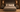 A rustic gray wooden bed frame with a high panel headboard and footboard, styled with beige and brown bedding, shams, and a textured throw blanket, set in a dark wood bedroom with matching nightstands and wall sconces.